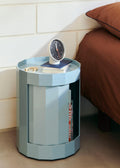 HAY Table Clock, Black, black dial table clock, on a pale blue round cabinet beside a bed with rust bedding and a white brick wall
