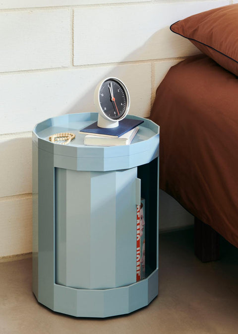 HAY Table Clock, Black, black dial table clock, on a pale blue round cabinet beside a bed with rust bedding and a white brick wall