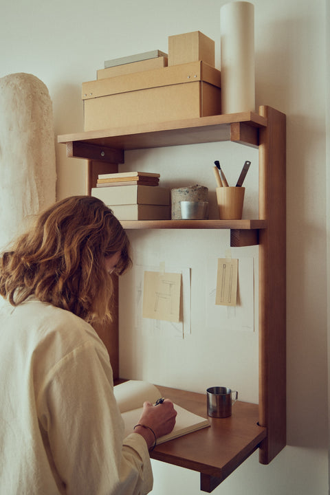 Form & Refine Beam Office Shelf 3, solid pine wall shelf with desk, person writing at the deep brown pine office shelf used as a wall desk