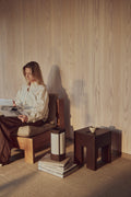 Form & Refine Monolith Side Table, solid wood side table, person reading in a wooden lounge chair beside the dark brown ash side table