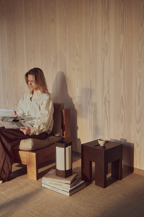 Form & Refine Monolith Side Table, solid wood side table, person reading in a wooden lounge chair beside the dark brown ash side table