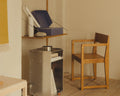 Frama Armrest Chair 01 in Warm Brown Birch, birch armchair beside a metal stove in a room with a yellow curtain and a shelf