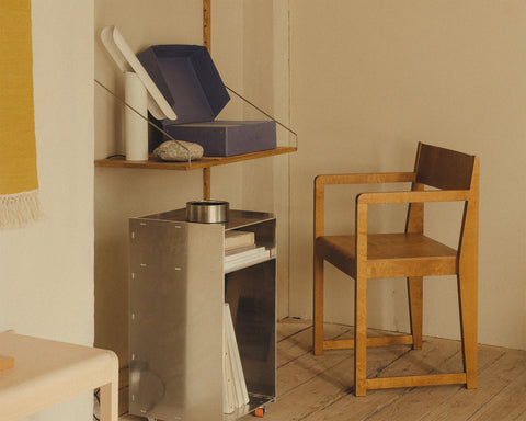 Frama Armrest Chair 01 in Warm Brown Birch, birch armchair beside a metal stove in a room with a yellow curtain and a shelf