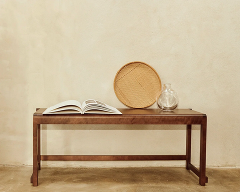 Frama Bench 01 in Dark Brown Birch, dark bench against a plaster wall holding an open book, a round woven tray and a glass carafe
