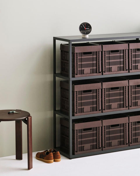 HAY Table Clock, Black, black dial table clock, on top of a black shelving unit of deep red crates, with a wood stool and shoes below