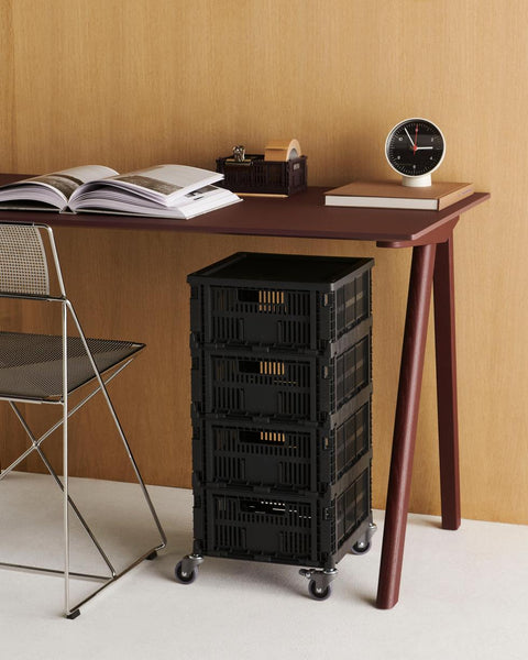 HAY Table Clock, Black, black dial table clock, on a wood desk with an open book, a chromed wire chair and charcoal crates on wheels