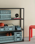 HAY Table Clock, Black, black dial table clock, on a black shelving unit of dusty blue crates, with a red stool and a basketball