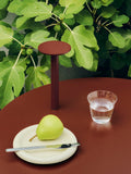 HAY Barro Plates, Ø24, Off-White, glazed terracotta plate, holding a pear on a dark red table with a glass and a red lamp