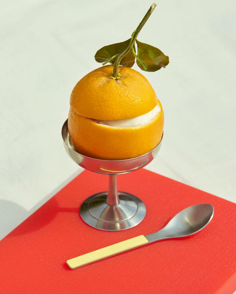 HAY Italian Ice Cup, Stainless Steel, stainless steel ice cream cup, holding a whole orange on a red chopping board with a yellow spoon