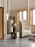 Audo Copenhagen Tearoom Lounge Chair, High Back, cream Tearoom Lounge Chair, High Back on a concrete floor, with a patterned lamp on a wooden table and a rattan screen
