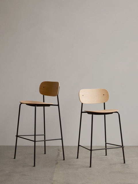 Audo Copenhagen Co Bar Chair, Non-Upholstered, Co Bar Chair and Co Counter Chair with natural oak veneer seats and black steel frames standing against a plain grey wall