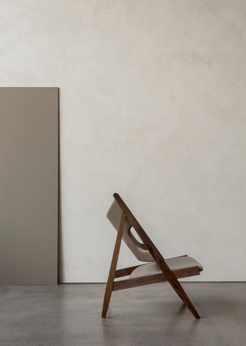 Side view of the Knitting Lounge Chair in oak with leather upholstery against textured plaster walls.