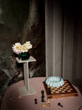 Museum Pedestal by String Furniture in beige holding a green glass vase of flowers, styled on a striped tablecloth beside a chessboard
