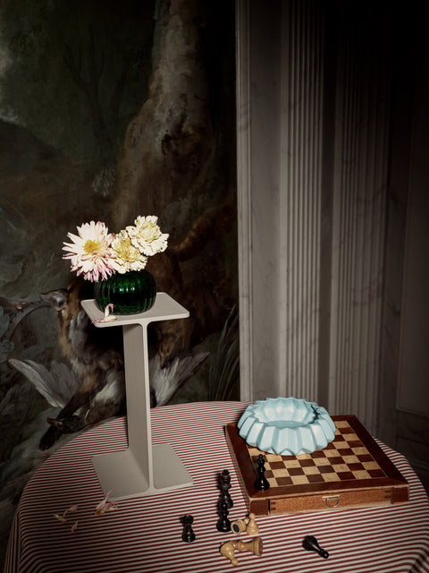 Museum Pedestal by String Furniture in beige holding a green glass vase of flowers, styled on a striped tablecloth beside a chessboard