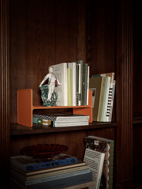 Museum Pedestal by String Furniture in orange turned on its side as a book stand in a wooden bookcase, holding books and a porcelain figurine