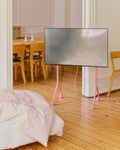 Pedestal Moon Light TV Stand in Bubble Gum, compact steel TV stand on wheels, holding a screen in a room with a wooden floor and a dining table behind