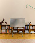 Pedestal Moon Light TV Stand in Mossy Green, compact steel TV stand on wheels, holding a screen in a room above a low bookshelf