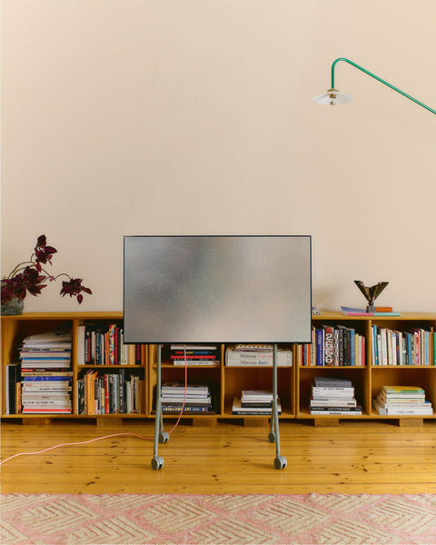 Pedestal Moon Light TV Stand in Mossy Green, compact steel TV stand on wheels, holding a screen in a room above a low bookshelf
