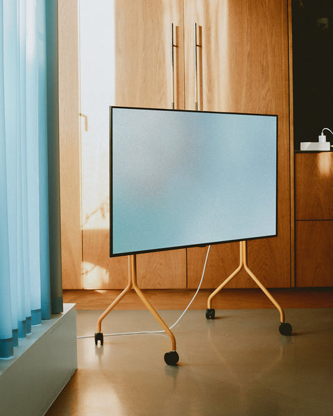Pedestal Moon Tall TV Stand in Apricot, holding a flat screen beside wooden cabinetry and a blue curtain