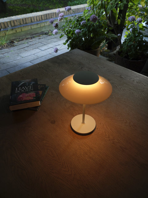 PH 80 Portable Lamp in white illuminated on a wooden table beside books with garden view through window