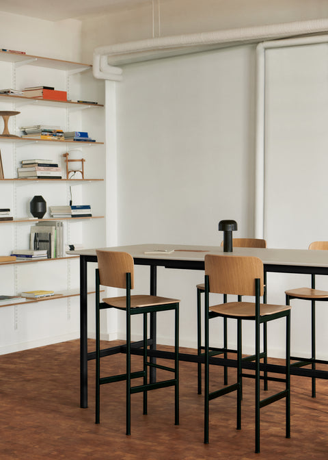 PLAN Barstools in oak and black arranged around a high table in a warm, Scandinavian-inspired room with wall shelving.