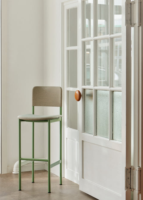 PLAN Barstool in modernist green upholstery placed beside a white-paneled door in a Scandinavian-inspired interior.