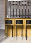 Black PLAN Barstools against a yellow bar with exposed metal fixtures and white tiled wall in a modern bar.