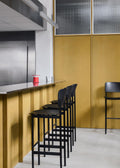 Black PLAN Barstools lined up along a stainless-steel bar counter with yellow paneling in a modern restaurant.