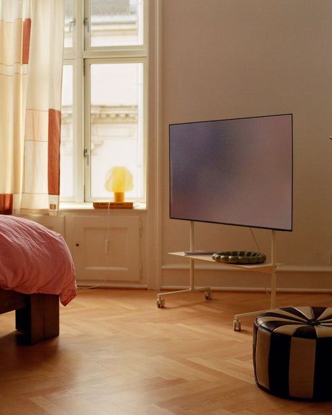 Pedestal Rover TV Stand in Pearl, holding a flat screen in a bedroom with a window, curtains and a striped pouf