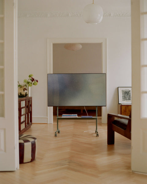Pedestal Rover TV Stand in Mossy Green, holding a flat screen in a room with a herringbone floor and a pendant lamp