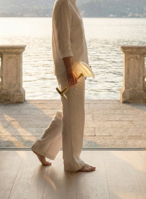 Woman in white linen carrying the lit Louis Poulsen PH 1/1 Portable lamp in brass across a lakeside terrace