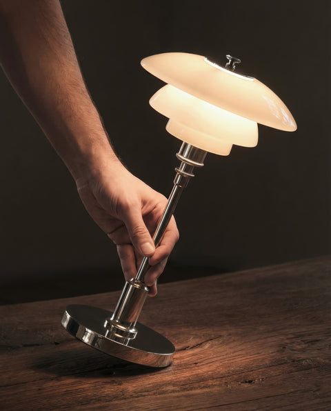 Hand placing the lit Louis Poulsen PH 1/1 Portable lamp in chrome on a wooden surface