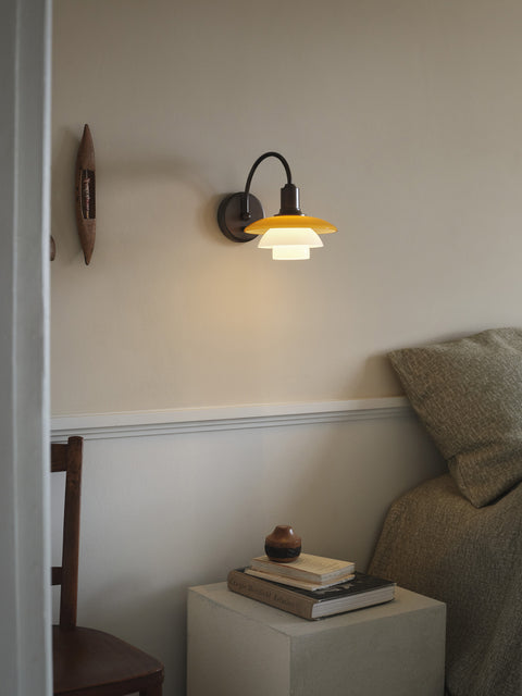 Louis Poulsen PH 2/2 Wall Lamp Centenary Edition lit above a bedside table stacked with books, beside a bed