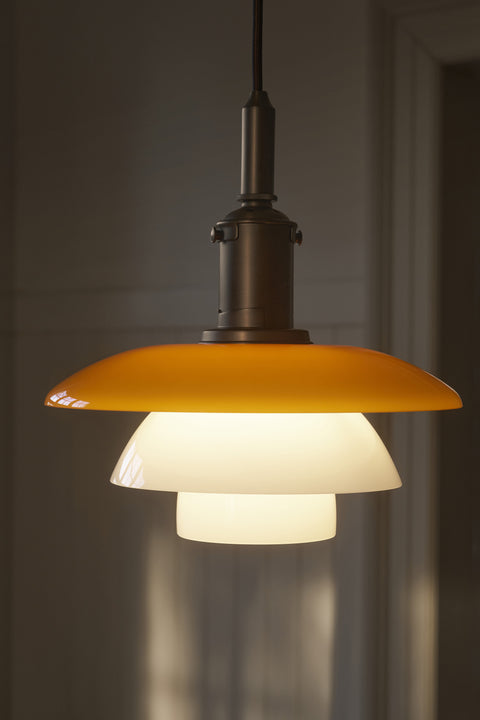 Louis Poulsen PH 3/3 Pendant Centenary Edition lit in a darkened interior, showing the amber upper shade and aged brass shade holder
