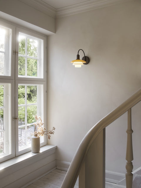 Louis Poulsen PH 2/2 Wall Lamp Centenary Edition lit on a stair landing wall beside a window overlooking a garden