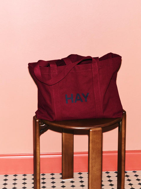 HAY Shopper Bag, Burgundy, cotton tote bag, burgundy shopper on a dark wood stool by a pink wall above chequered tiles