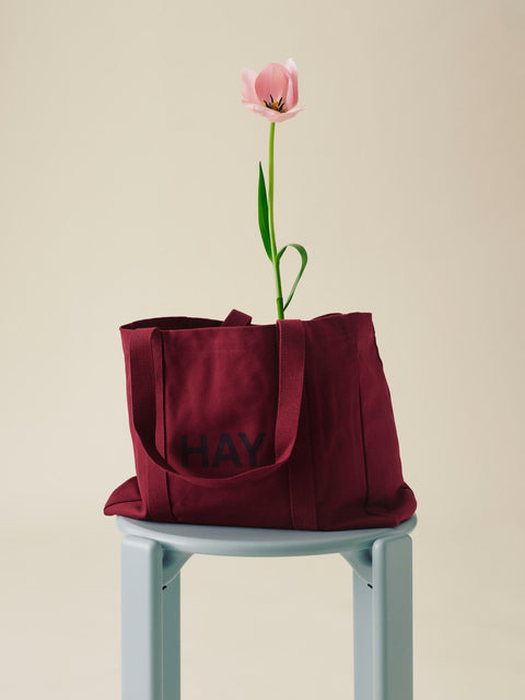 HAY Shopper Bag, Burgundy, cotton tote bag, burgundy shopper on a pale blue stool with a pink tulip stem rising from it