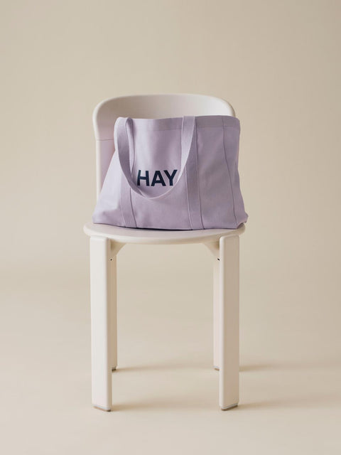 HAY Shopper Bag, Lavender, cotton tote bag, pale lilac shopper resting on a white wooden chair against a warm beige wall