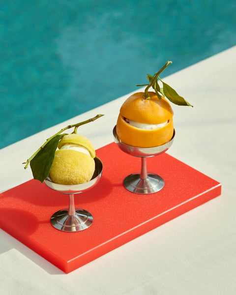 HAY Italian Ice Cup, Stainless Steel, stainless steel ice cream cup, two cups holding an orange and a lemon on a red board by turquoise water