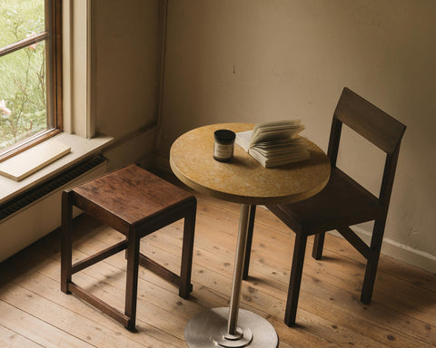 Frama Table 57 in Yellow Limestone, speckled stone top holding a jar and folded papers by a window, wooden stool and dark chair nearby