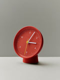 HAY Table Clock, Red, red table clock, angled on a white surface against a soft grey green backdrop, casting a long shadow
