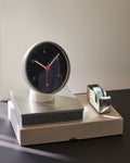 HAY Table Clock, Black, black dial table clock, white cased clock on a grey tray and box beside a tape dispenser on a dark desk