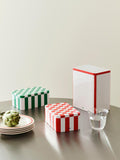 HAY Tin Container, Large, Off-White and Red, lidded tinplate storage tin, on a steel table with two smaller tins, a water glass and an artichoke