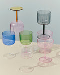 HAY Tint Wine Glasses, Blue, tinted borosilicate wine glasses, blue, green, pink and smoke glasses stacked on a cream table, pale blue backdrop