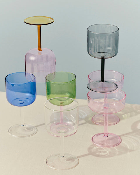 HAY Tint Wine Glasses, Blue, tinted borosilicate wine glasses, blue, green, pink and smoke glasses stacked on a cream table, pale blue backdrop