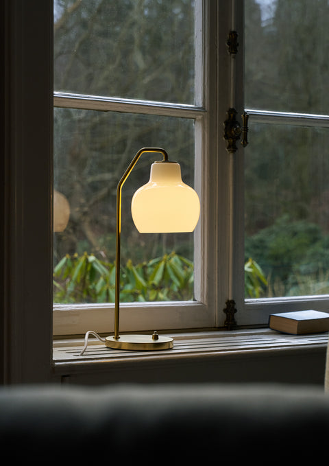 Louis Poulsen VL Ring Crown table lamp on a window sill with brass base and opal glass shade, modern Scandinavian lighting.