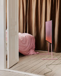 Pedestal Viva TV Stand in Bubble Gum, steel TV floor stand, holding a screen in a bedroom with curtains and pink bedding
