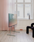 Pedestal Viva TV Stand in Mossy Green, steel TV floor stand, holding a screen in a room with pink curtains and a dark armchair