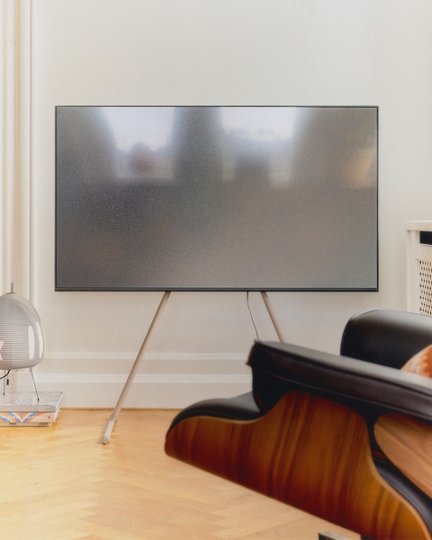 Pedestal Viva TV Stand in Oat, steel TV floor stand, holding a screen in a room with a wooden floor and a lounge chair