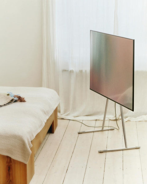 Pedestal Viva TV Stand in Chrome, steel TV floor stand, holding a screen in a bedroom beside a bed and a window